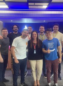 #Paratodosverem: Foto de um grupo de oito pessoas posando juntas e sorrindo para a câmera em um ambiente interno com iluminação azul no teto e um fundo escuro com um letreiro luminoso. Todos estão vestidos de forma casual, alguns usando crachás. Uma mulher ao centro veste uma blusa preta e calça bege, enquanto um homem à direita veste uma camiseta azul com os dizeres "Felicidade, Inclusão, Crédito & Inovação" e faz um sinal de positivo com a mão. Os demais integrantes usam camisetas em tons neutros, alguns com detalhes coloridos. O clima da foto é descontraído e de camaradagem.
